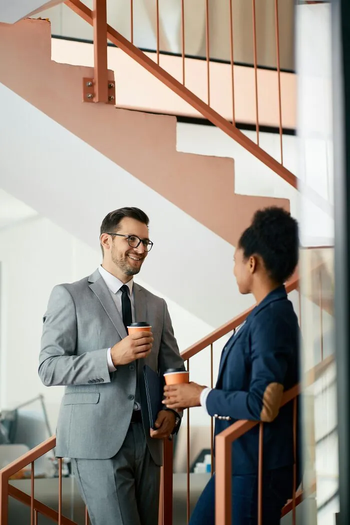 Two colleagues chatting on an office staircase with coffee cups, discussing weird at work stories.