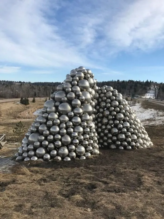 A large pile of metallic silver spheres forming two mounds in an open field, showcasing outdoor public art.