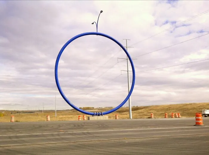 A huge blue metal ring on a concrete base next to a road under a cloudy sky. Bizarre public art installation.