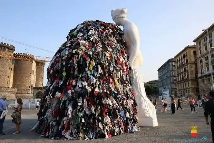 A large public art installation featuring a white statue standing next to a huge pile of clothes