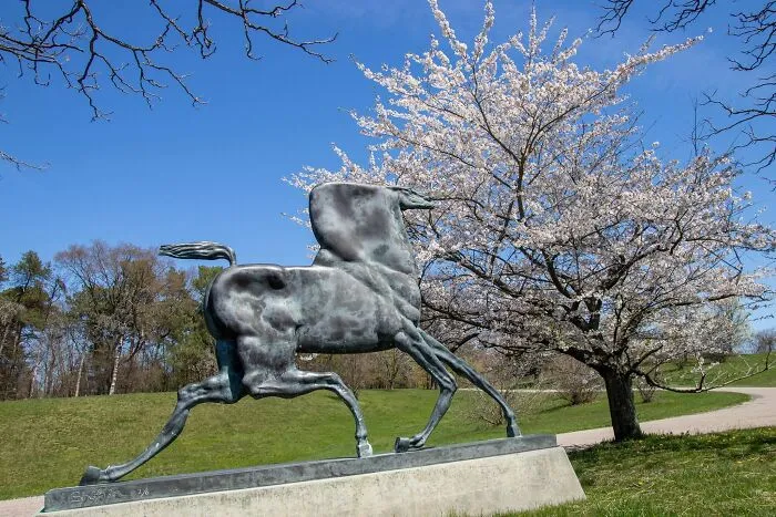 A bizarre public art sculpture of a running horse in a grassy park next to a blooming cherry blossom tree