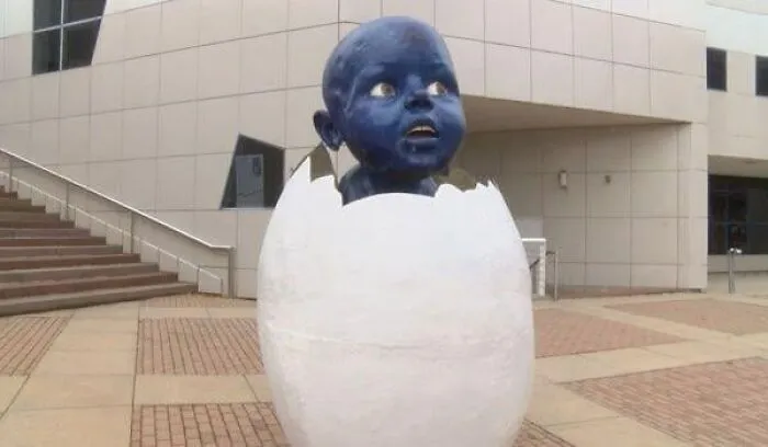 A creepy public art statue of a blue baby hatching from a white egg shell