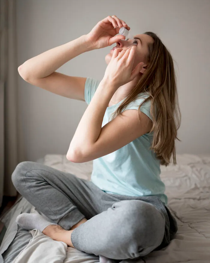 Young woman applying eye drops while sitting on a bed, addressing common Gen Z health problems.