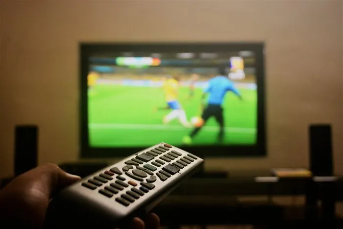 Hand holding remote control toward TV showing soccer match