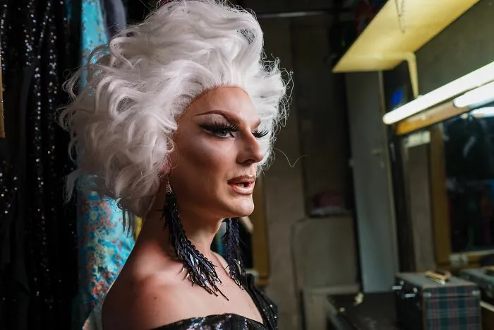 Person in drag makeup and white wig looking in dressing room mirror