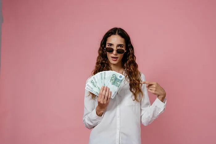A woman in a white shirt and sunglasses holding a fan of euro bills, showing people behaving like they are owed everything