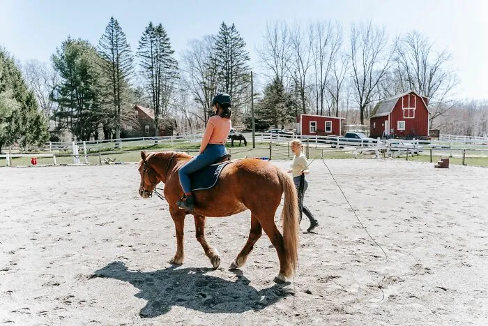 A young woman riding a brown horse in an outdoor arena while a trainer guides her, depicting entitled behavior themes