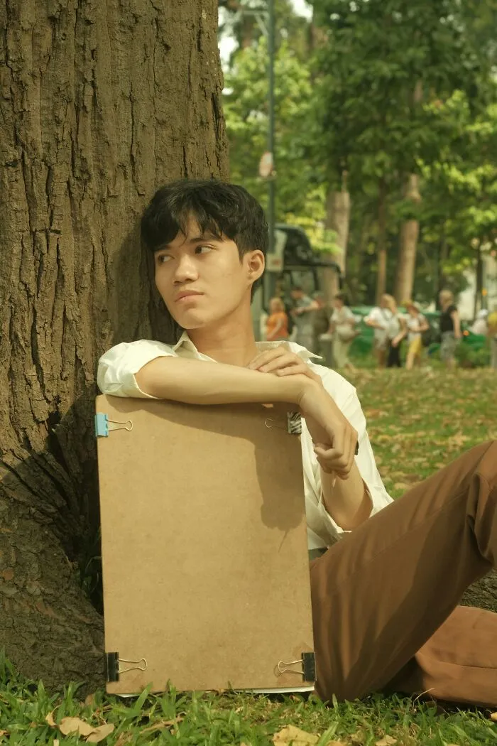 Young man sitting against tree holding drawing board outdoors