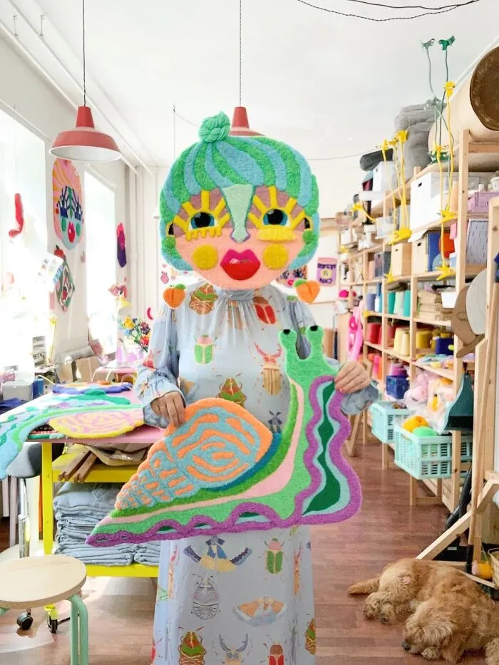 Artist Ina Dyreborg posing with her colorful handmade snail rug and character mask in her studio.