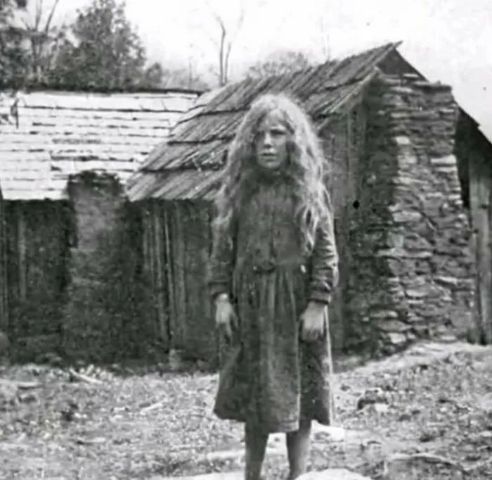 Black and white photo of a girl with long hair standing in front of rustic wooden and stone buildings