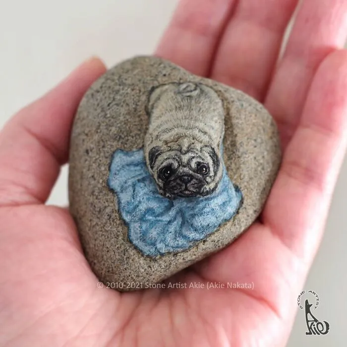 Hand holding stone painted as a detailed pug dog with blue collar art