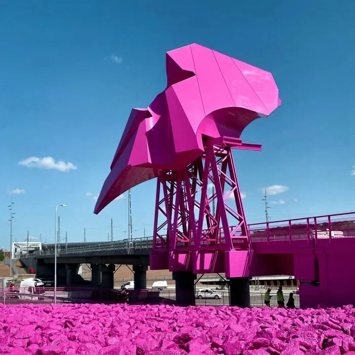 A bright pink public art sculpture integrated into a bridge over a rocky ground painted entirely in pink.