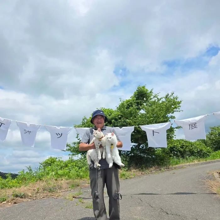 Japanese grandpa smiling outdoor photoshoot holding his two white cats in front of hanging shirts.