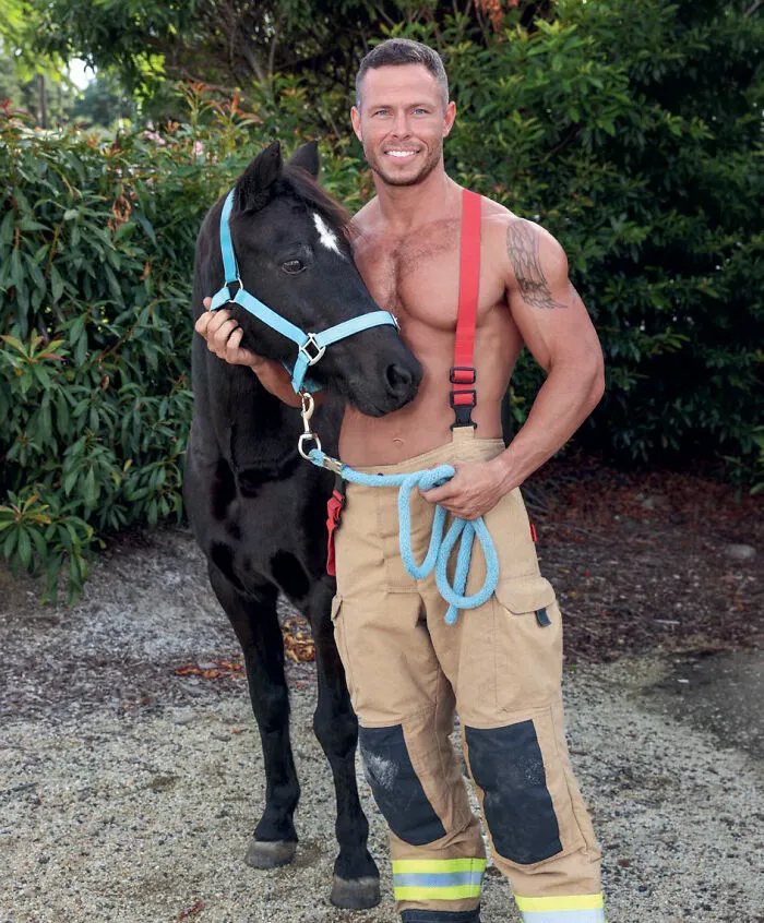 Shirtless firefighter with red suspenders holding black horse with blue halter outdoors
