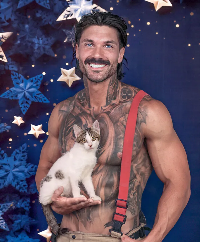Muscular firefighter with tattoos holding a white spotted kitten with starry background