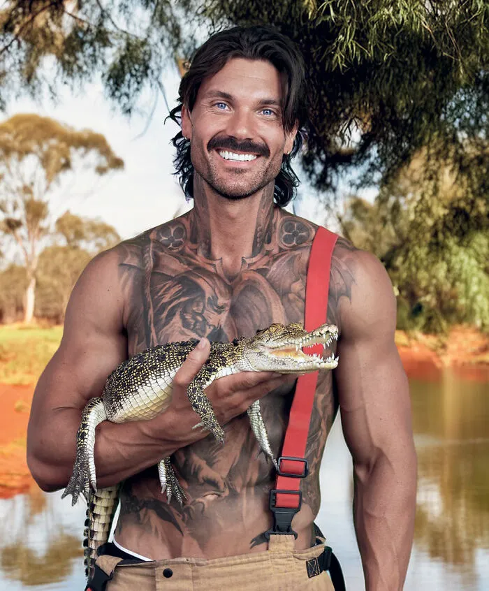 Muscular Australian firefighter with tattoos holding a young crocodile by water