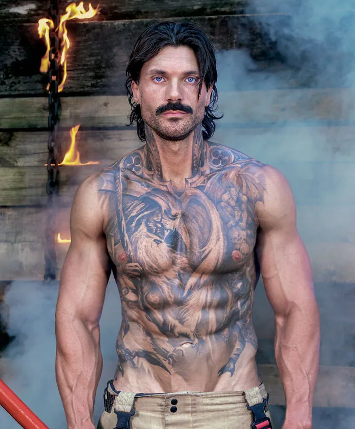 Tattooed firefighter with dark hair and mustache standing amid flames and smoke