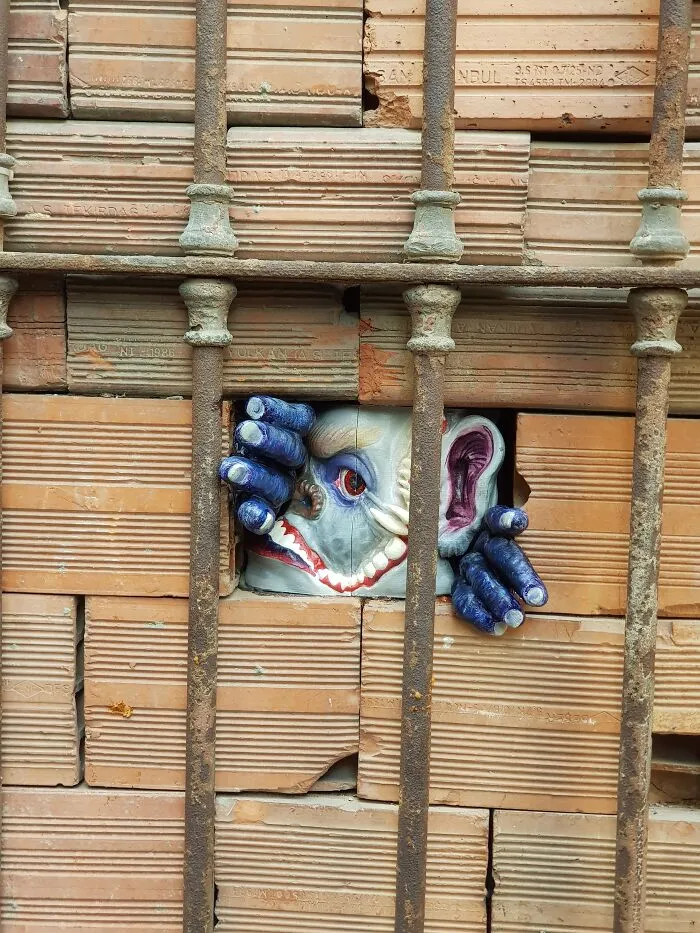 Creative art collective monster figure peeking through gaps in a brick wall behind iron bars.