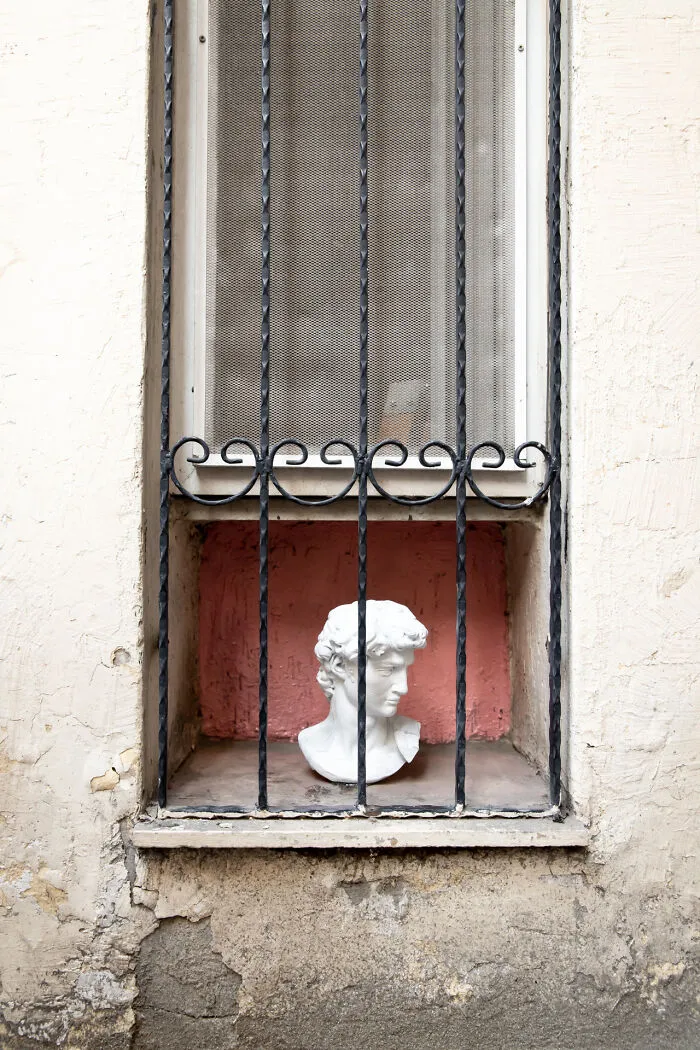 Creative art collective marble bust sculpture behind black iron window grates on a pink background.