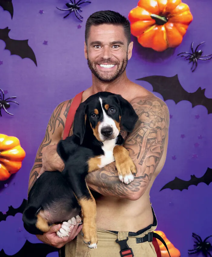 Tattooed Australian firefighter holding a puppy with Halloween-themed backdrop for 2027 firefighter calendar