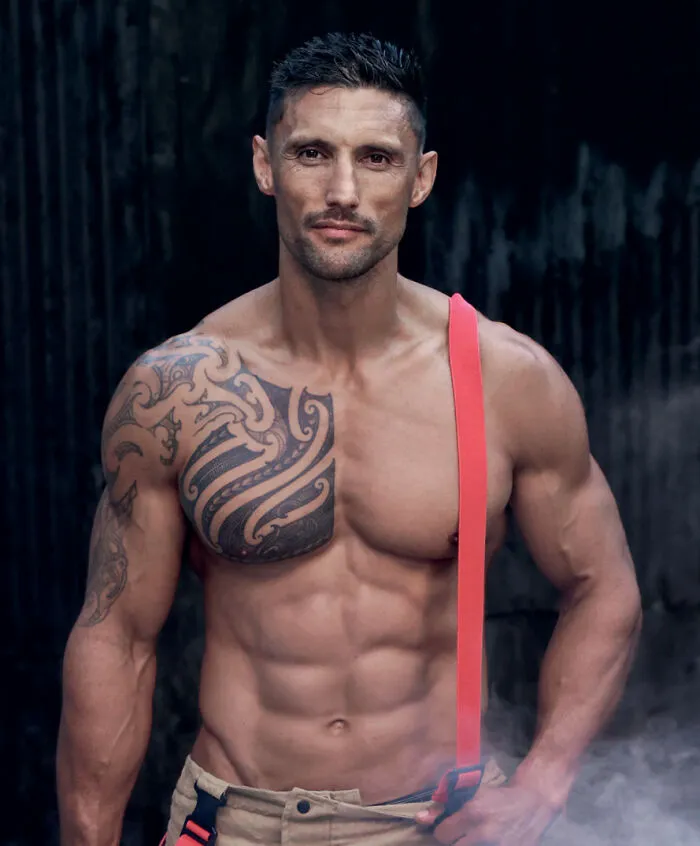 Muscular firefighter with tattoos and red suspenders posing against dark background