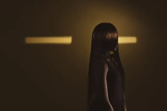 A young woman standing in low light with a beam of light behind her, highlighting human expression in portrait photos.