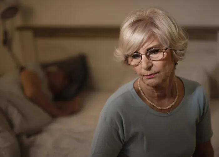 A worried senior woman sitting on the edge of a bed, reflecting dramatic mil stories.