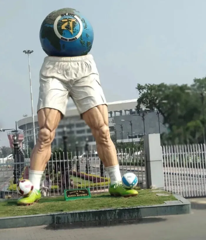A surreal public art sculpture of muscular legs with soccer balls and a globe torso outside a stadium