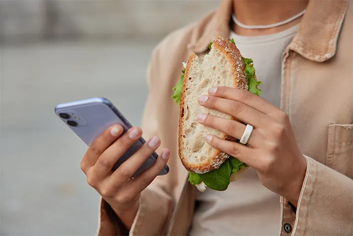 Close-up of a person holding a sandwich and typing on a smartphone during a break from work.