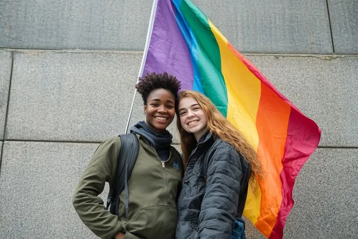 Two happy young women smiling with a rainbow pride flag behind them.