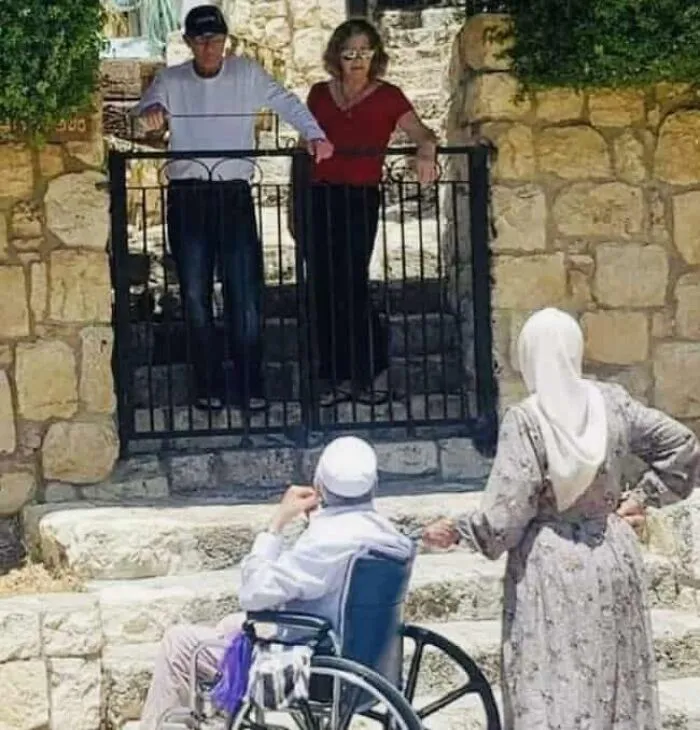 A man in a wheelchair and a woman stand below a gate, illustrating barriers in modern life in America.