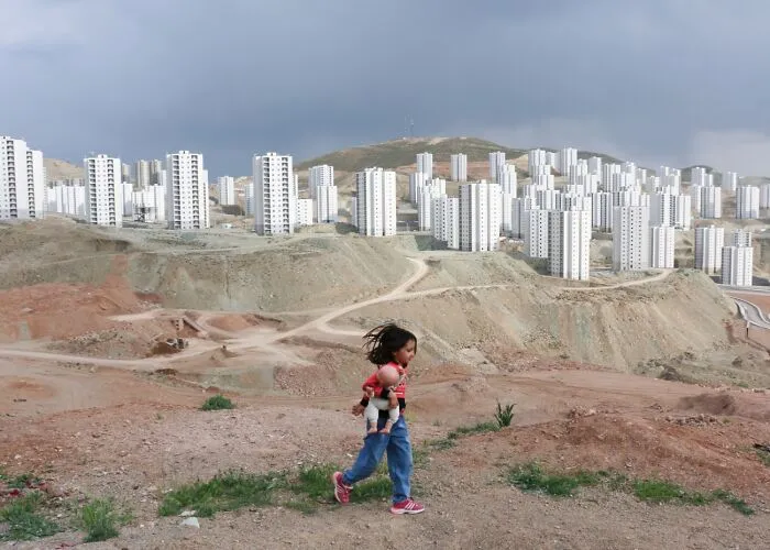 Child holding doll walking near new city buildings, best pics Siena International Photo Awards 2026
