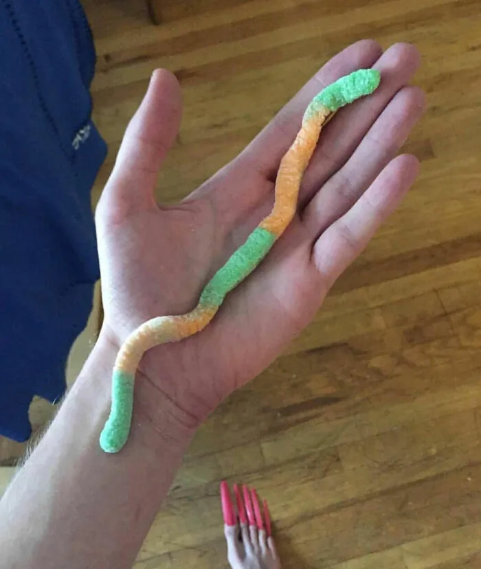 Close up of a long gummy worm on a hand, with a funny background detail of toes with long pink nails.