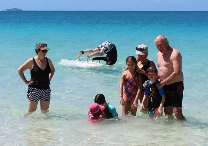 A surfer wipes out and hijacks a family photo in the background at a tropical beach.