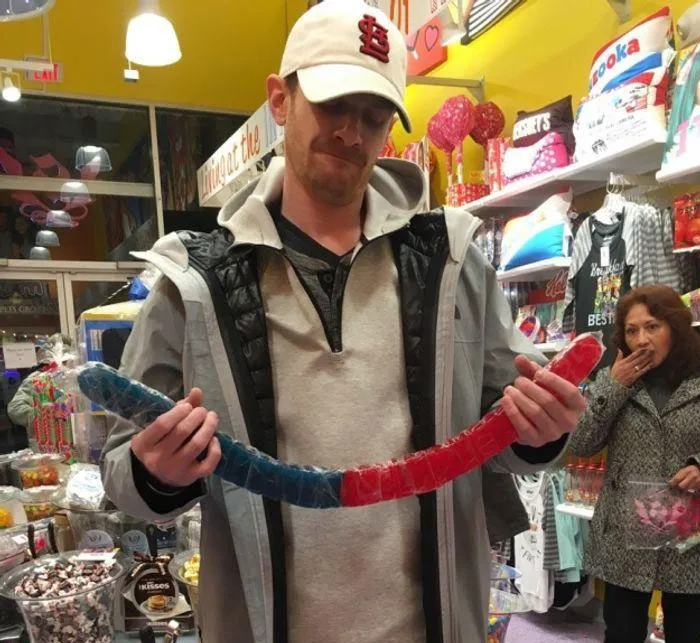 A shocked woman in the background of a man holding a giant gummy worm.