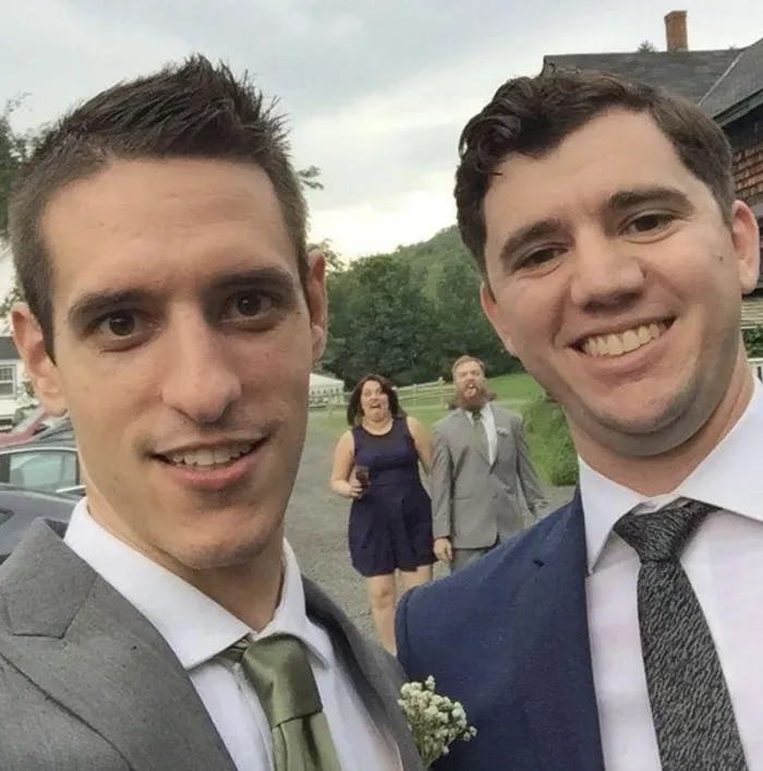 Funny photobomb of a couple making faces in the background behind two smiling men taking a selfie.