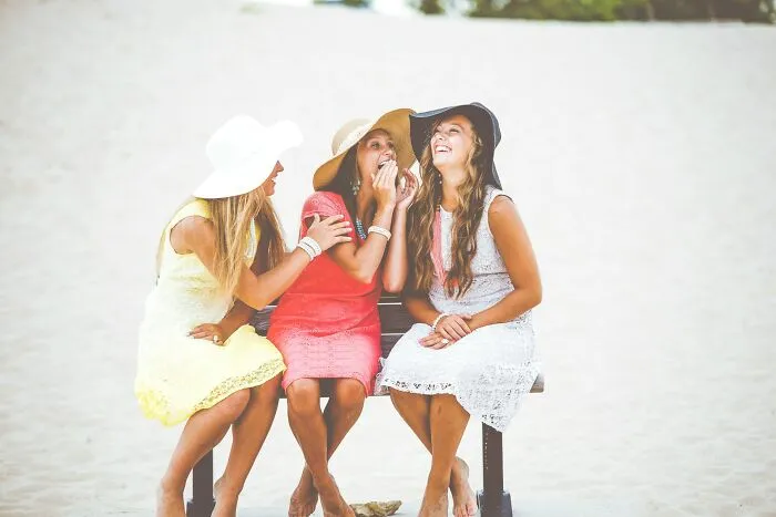 Three young women in sun hats sitting on a beach bench laughing together at funny instances.