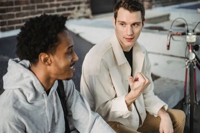 Two friends sitting outside talking near a bicycle, casually sharing intelligent insults