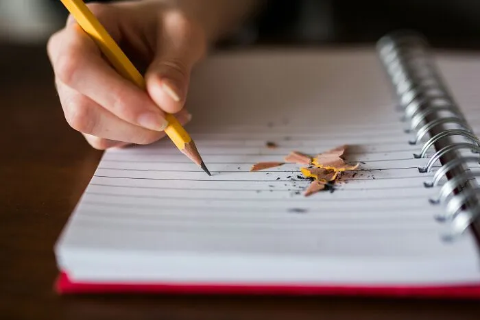 A hand writing with a yellow pencil in a notebook, taking note of intelligent insults.