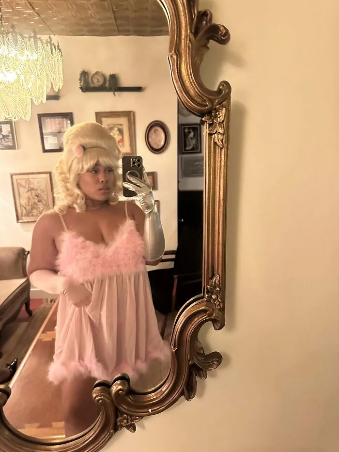 Woman posing in a mirror wearing a glamourous pink fluffy Halloween costume with blonde styled wig.