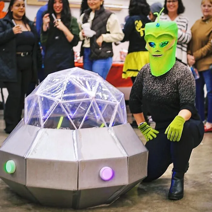 A person in a green alien mask kneeling next to a spaceship display for next level Halloween costumes