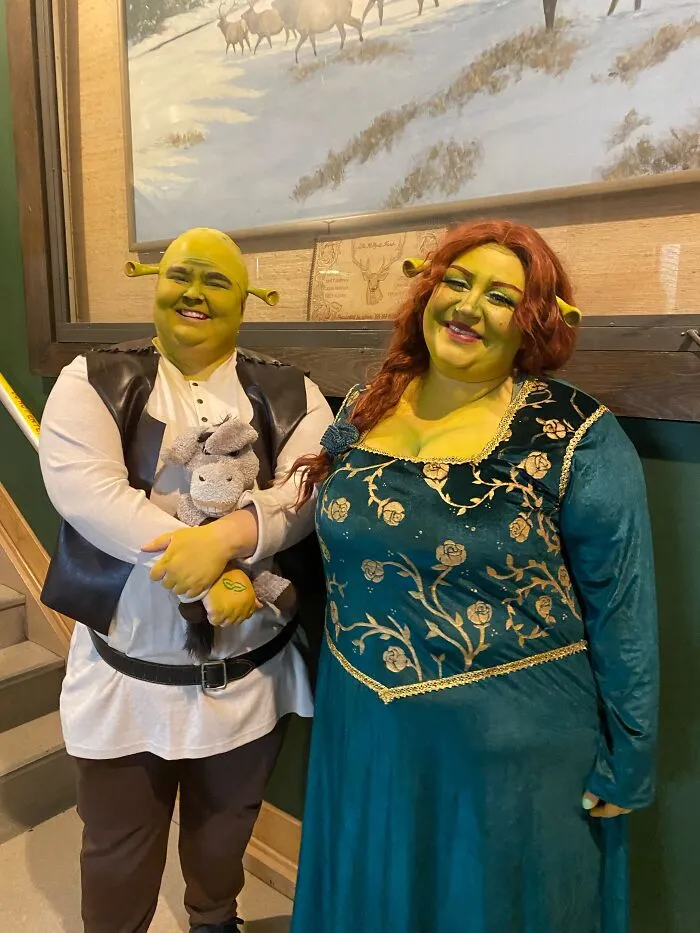 Two adults dressed up in detailed Shrek and Fiona couple Halloween costumes standing indoors.