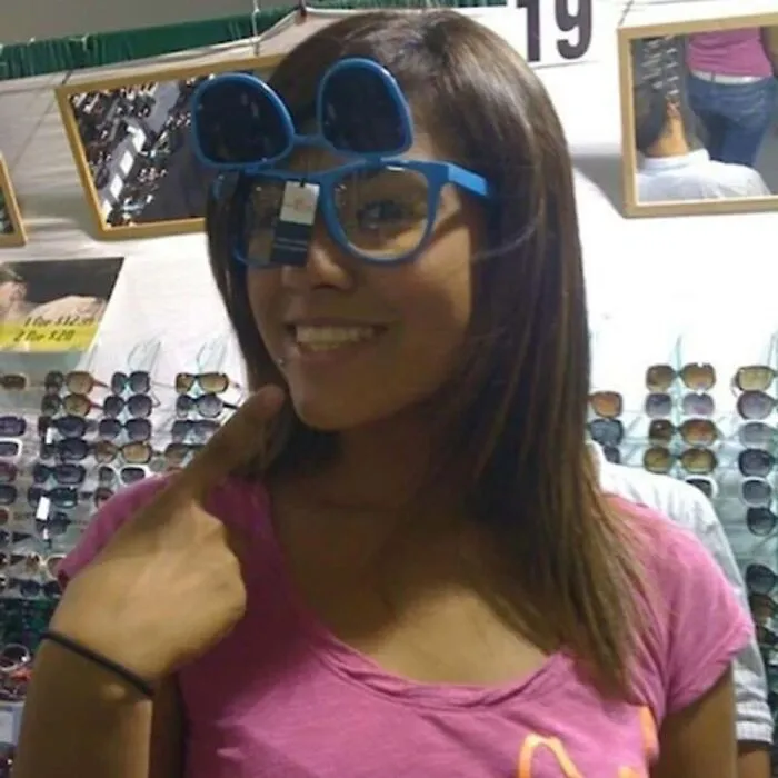 A woman smiling wearing novel blue sunglasses in a funny mirror pic setup at a store.