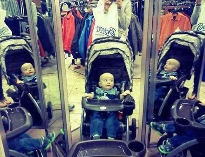Baby in stroller looking shocked at his mirror reflection in a clothing store.