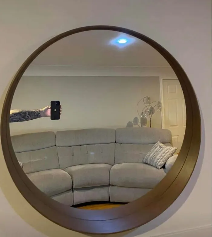 Tattooed arm holding a smartphone extending into frame to take a mirror pic of a round wall mirror over a sofa.
