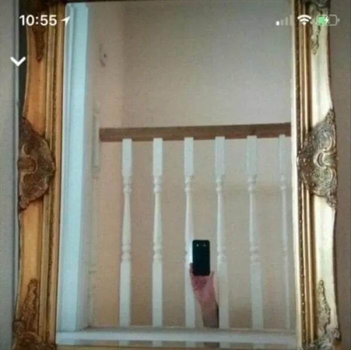 A hand holding a smartphone appearing from behind white stair banisters captured in an ornate mirror pic.