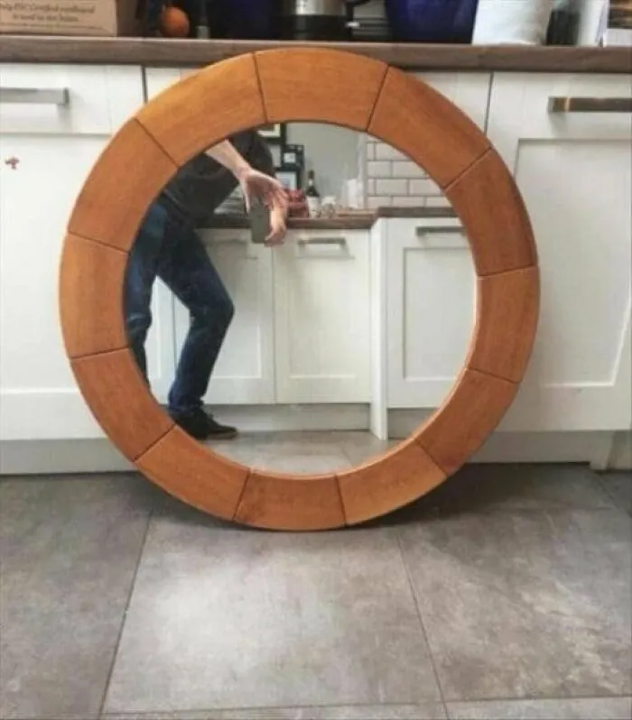 Person bending sideways to take a photo of a large round wooden framed mirror pic resting against kitchen cabinets.