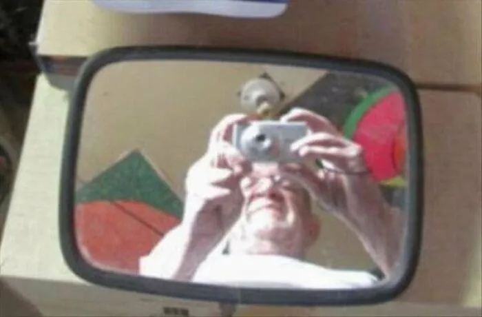 Man taking a selfie with an old silver digital camera reflected in a small black frame mirror pic.