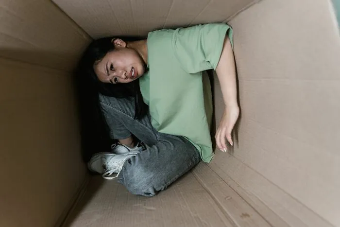 A young woman curled up inside a tight cardboard box, illustrating claustrophobia in crazy Airbnb stories.