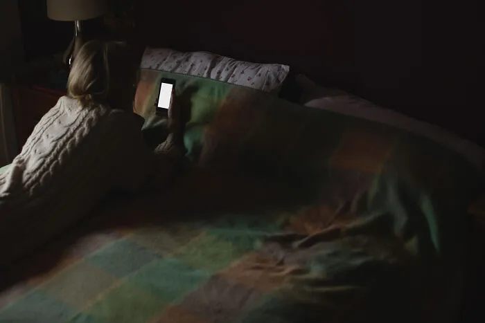 Person lying in bed looking at a smartphone screen in a dim room with spooky vibes